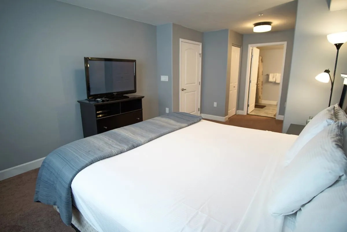 A spacious master bedroom featuring a large bed with crisp linens, a nightstand with lamp, and a television mounted opposite the bed. There is a doorway leading directly to the bathroom, suggesting en suite convenience and privacy. The subtle, serene color palette and tasteful decor contribute to a restful atmosphere.