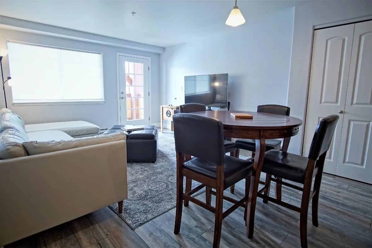 A spacious interior featuring a plush leather sectional sofa, a large dining table with ample seating, a wall-mounted HDTV, and large windows bathing the space in natural light. The scene captures both relaxing and dining activities, with views extending toward the covered deck and mountains beyond.