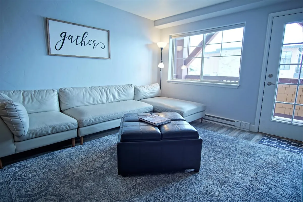 A welcoming living space with a leather sectional sofa, coffee table, and a large window letting in southern light. The 'gather' sign on the wall sets a tone of togetherness and relaxation, inviting board games or cozy evenings with loved ones.