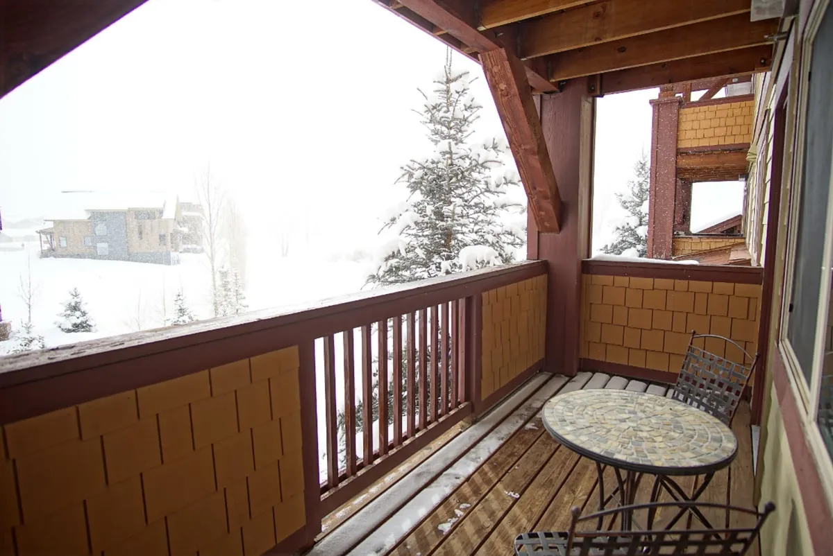 A tranquil covered deck with a small round table and metal chairs, overlooking snow-covered trees and framed by a wooden railing. The scene suggests a serene spot for morning coffee or enjoying alpenglow sunsets, connecting interior comfort with outdoor beauty.