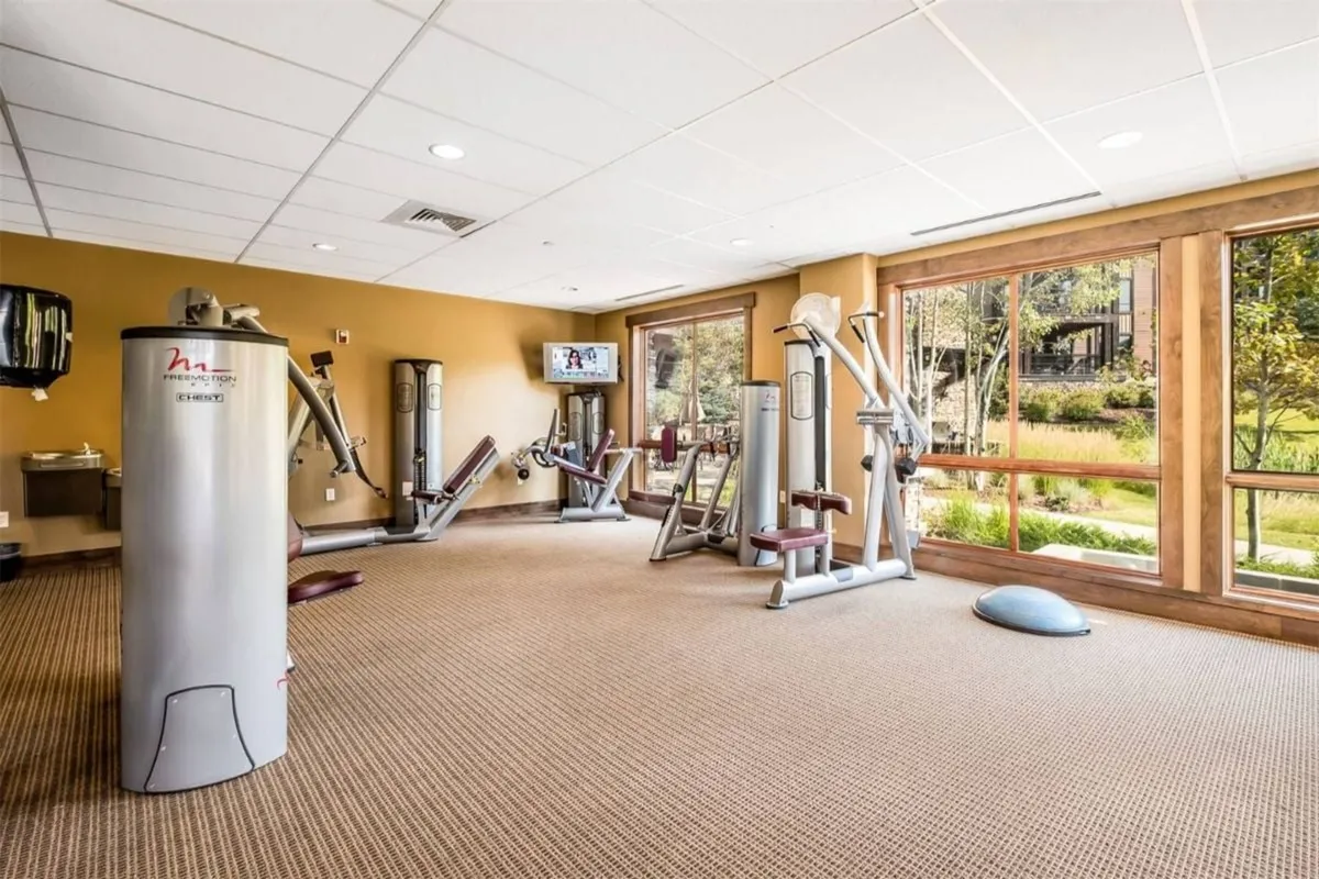 A modern fitness center with exercise machines lined up in front of large windows that let in natural light. The room is spacious and bright, creating an energetic atmosphere for workouts and wellness activities.