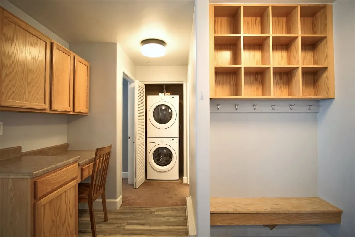 This image depicts a practical, well-organized utility space featuring a laundry area with a washer and dryer, wooden cabinets for storage, a built-in cubby and coat hooks for organizing outdoor gear, and a small desk with a chair. The area is tidy and functional, clearly designed for convenience and efficient storage.