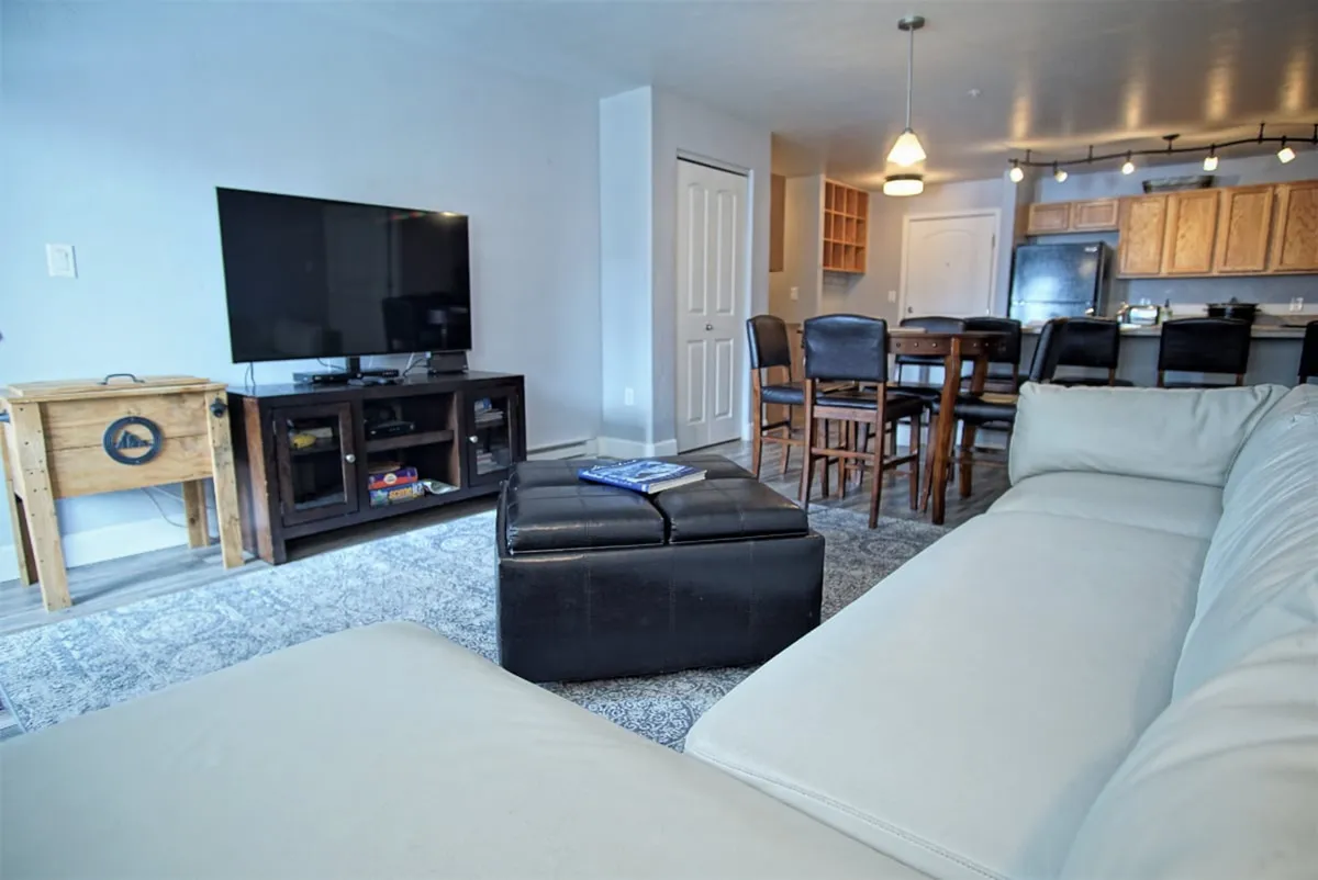 An open-plan living and dining area features a white sectional sofa, a dark coffee table, and a dining area with bar stools. The scene is set for entertaining, sharing meals, and group activities, with plenty of space for guests to gather.