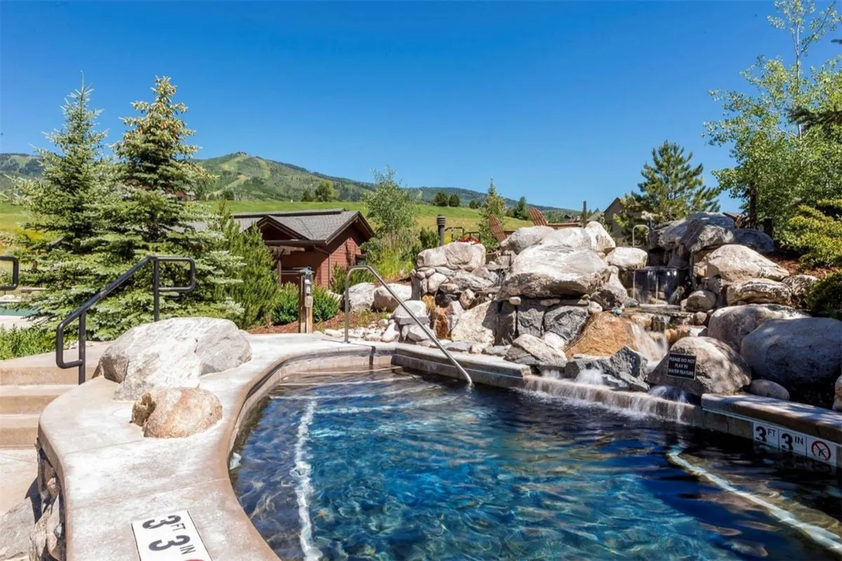 A tranquil exterior scene featuring a large outdoor hot tub with a waterfall, surrounded by lush greenery and mountain views. The inviting water and relaxed atmosphere hint at year-round enjoyment.
