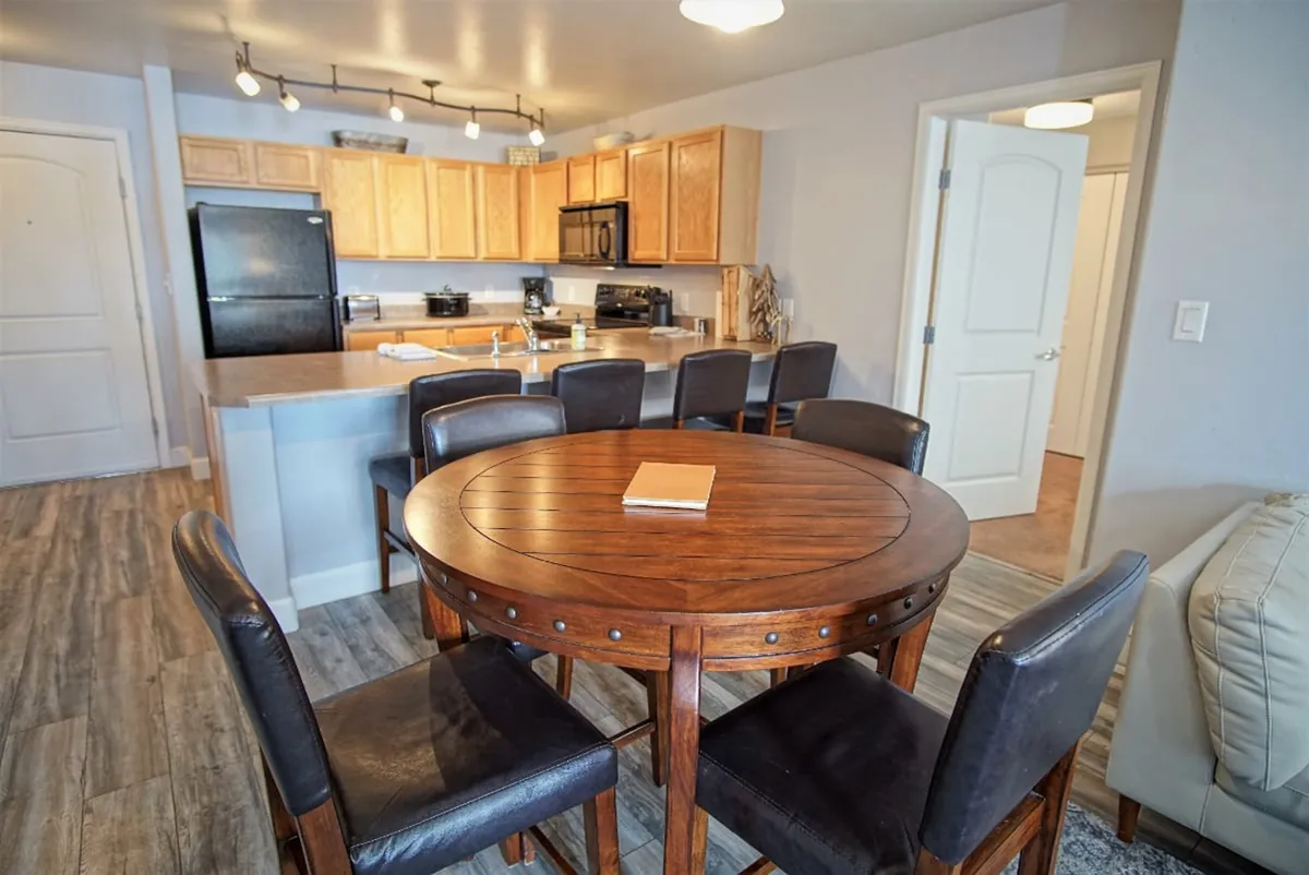 A cozy dining area with a round wooden table surrounded by chairs, seamlessly integrated with the open kitchen. The kitchen features black appliances, light wood cabinetry, and an open layout ideal for family meals and cooking together.